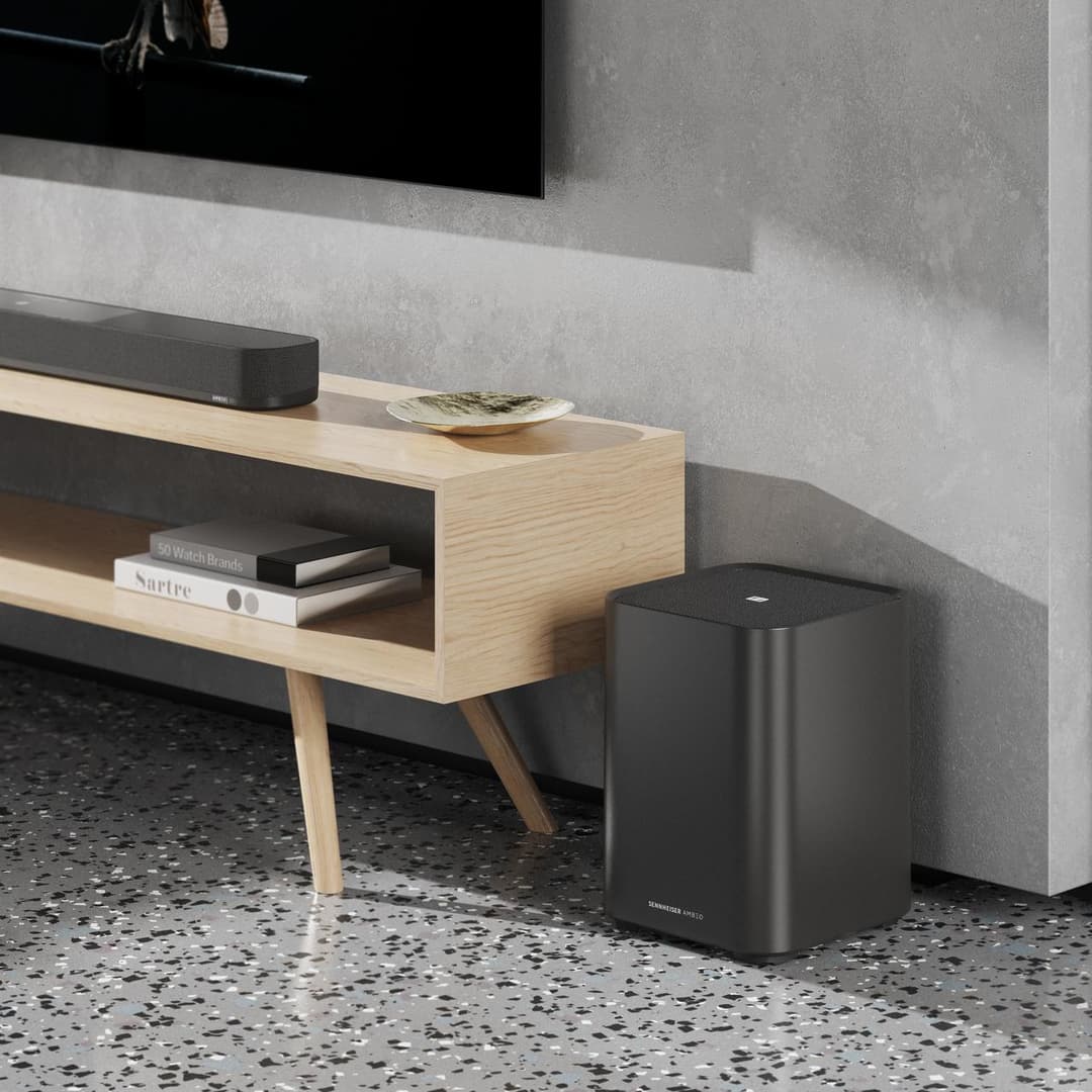 Barre de son Sennheiser AMBEO Soundbar Plus noire posée sous un meuble en bois clair, accompagnée d'un subwoofer noir, dans un salon moderne