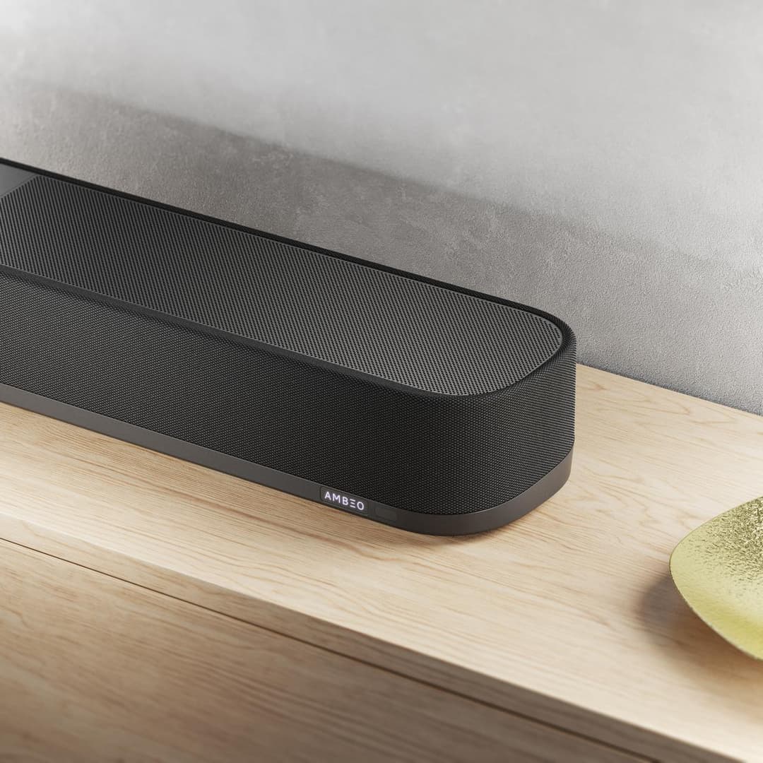 Barre de son Sennheiser AMBEO noire et compacte posée sur un meuble en bois clair devant un canapé gris