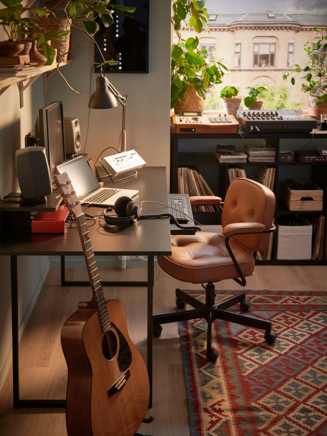 Studio de production musicale avec guitare acoustique, équipements audio, chaise en cuir marron et plantes vertes près d'une fenêtre