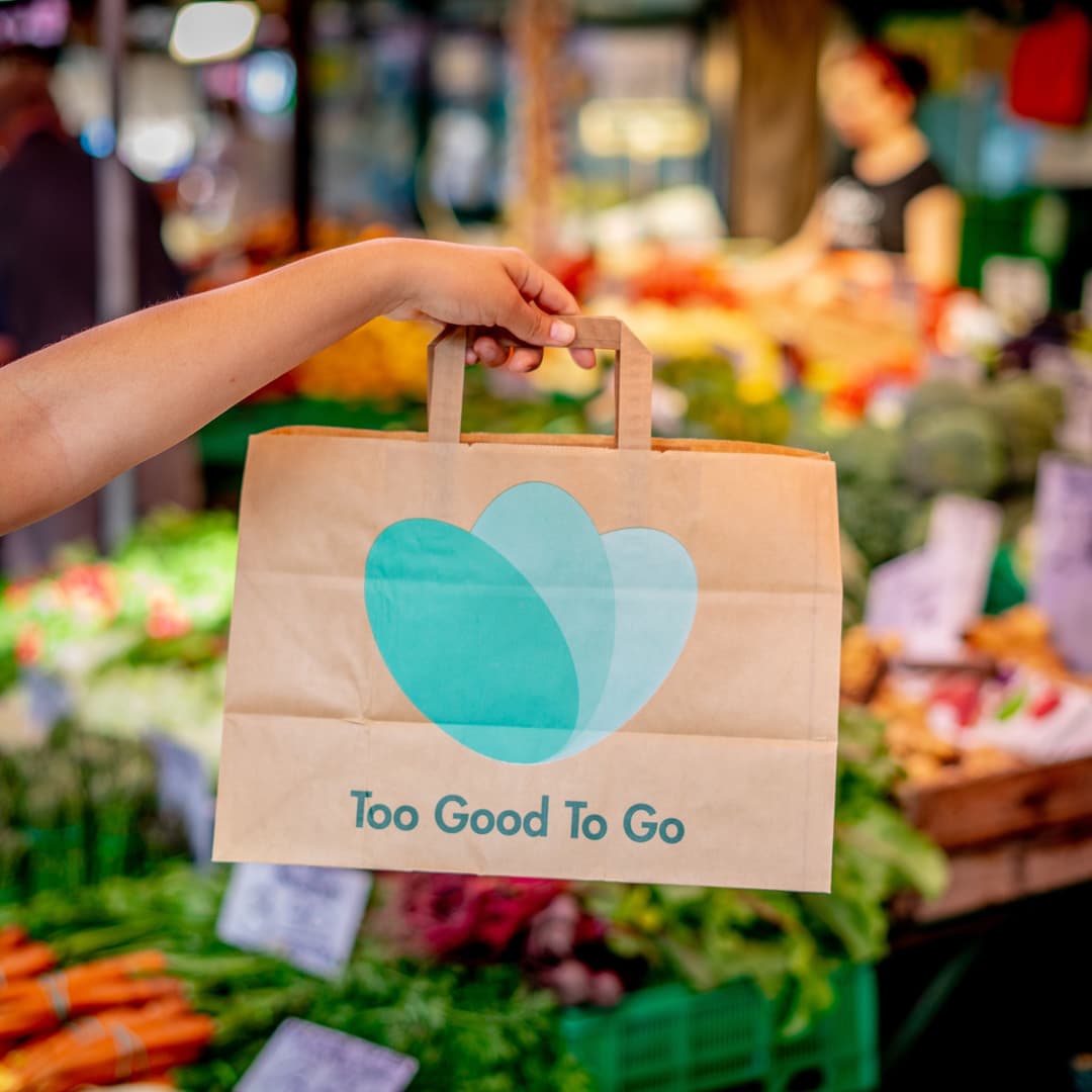 Sac en papier kraft avec logo Too Good To Go tenu à la main dans un marché avec fruits et légumes en arrière-plan