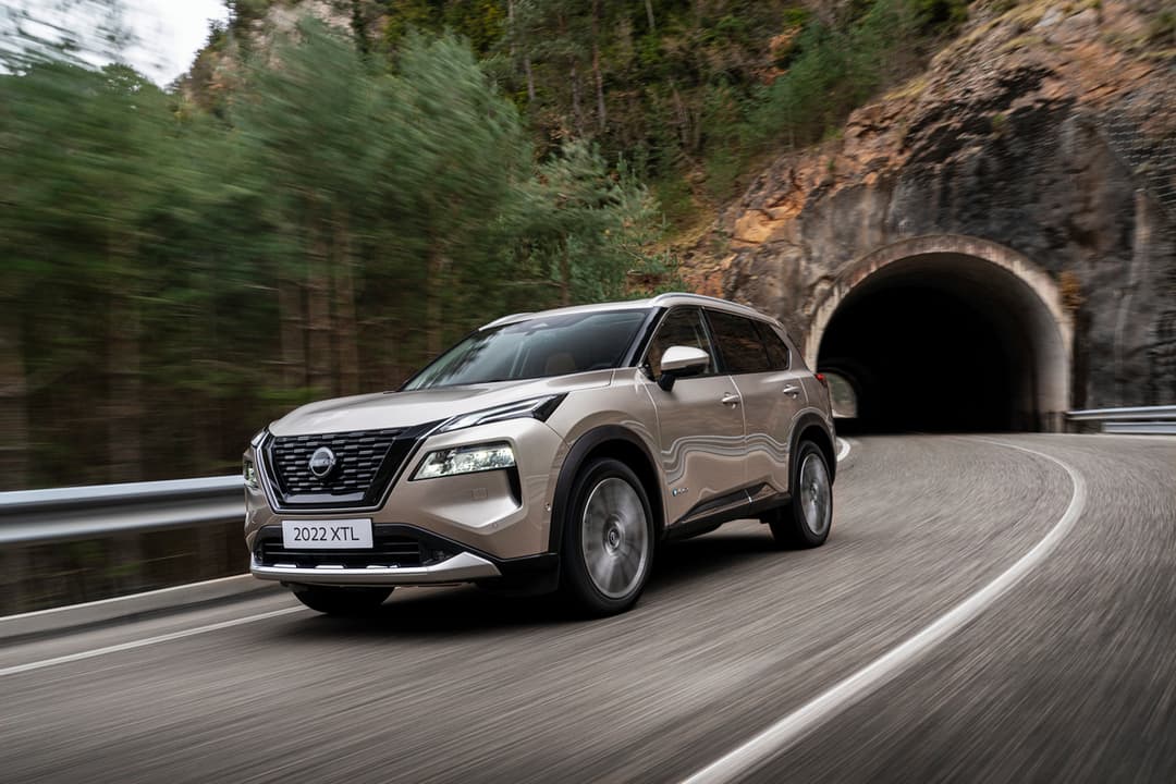 Nissan X-Trail beige métallisé roulant sur une route de montagne sinueuse avec tunnel rocheux en arrière-plan