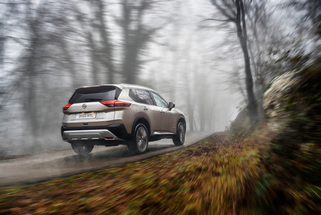 Nissan X-Trail beige en mouvement sur une route forestière sinueuse, aux arbres nus automne