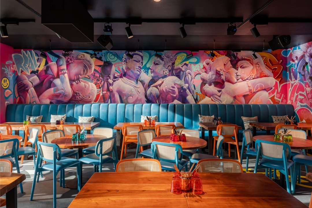Salle de restaurant colorée avec banquette turquoise, tables en bois, chaises orange et bleu, fresque murale street art aux teintes rose et orange