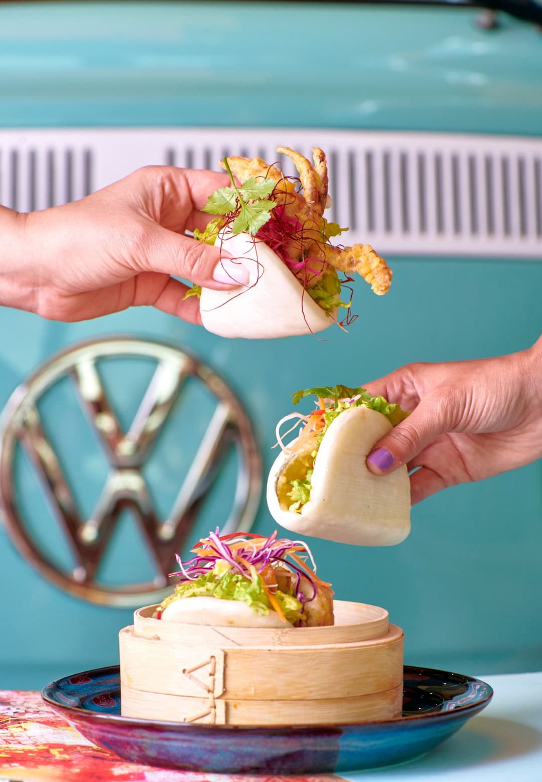 Bao et wraps garnis de laitue colorée et sauce, tenus en main devant un logo Volkswagen bleu