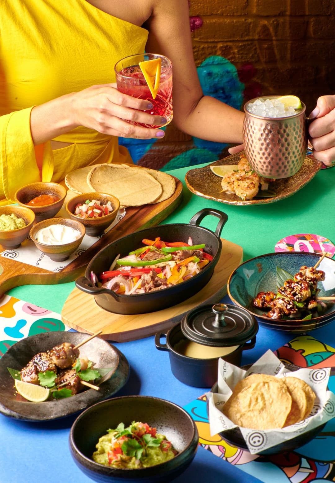 Table colorée avec street food variée et deux personnes tenant des cocktails colorés et une tasse métallisée