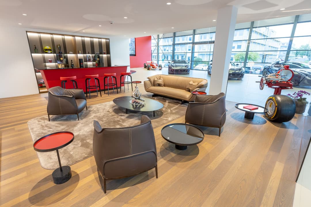 Showroom Ferrari rénové avec espace de détente moderne, fauteuils marron, bar rouge et véhicules visibles en arrière-plan