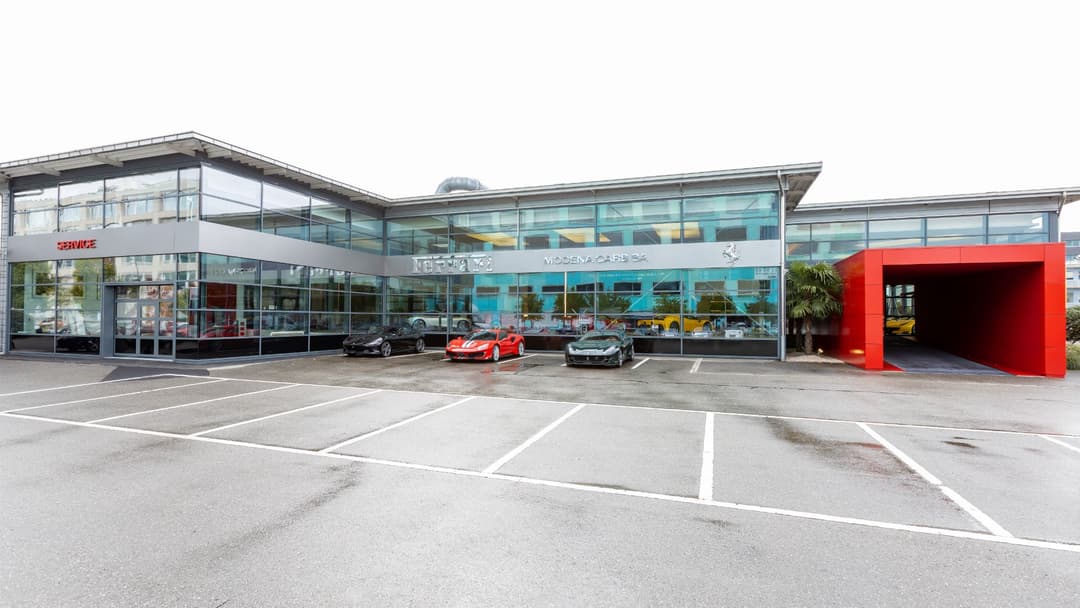 Showroom Modena Cars à Genève avec façade vitrée turquoise, entrée rouge et Ferrari rouge garée devant