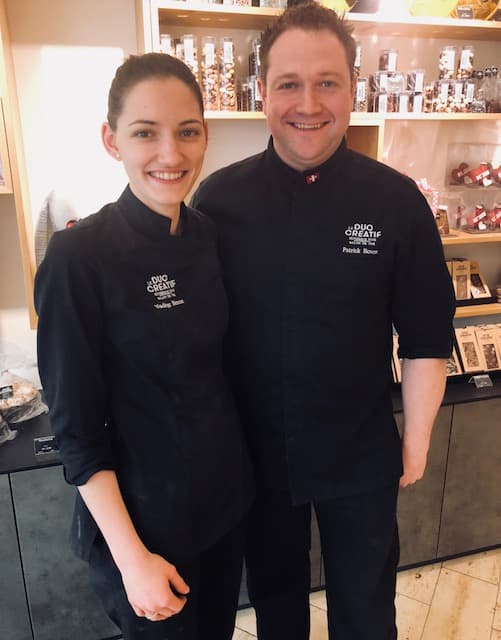 Nadège et Patrick, deux pâtissiers, posent souriants en tenues noires dans leur pâtisserie, avec des étagères de produits sucrés en arrière-plan
