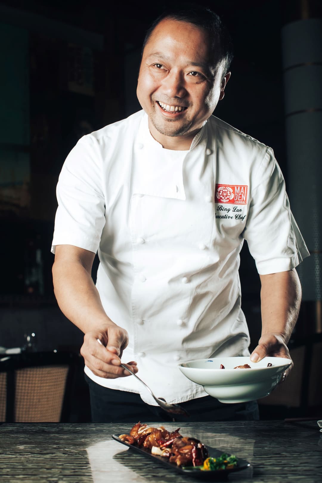 Le chef Luo Bing en tenue blanche sourit en tenant une assiette au-dessus d'un plat présenté sur le comptoir