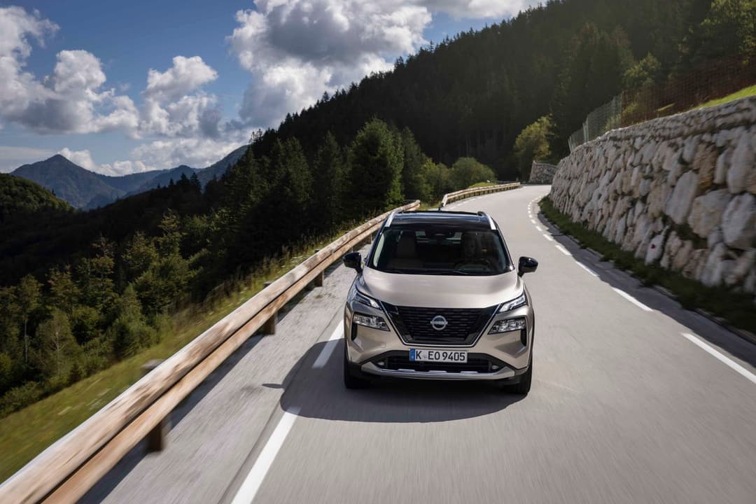 Nissan X-Trail blanc circulant sur une route sinueuse en montagne, entouré de forêts de sapins et de paysages alpins