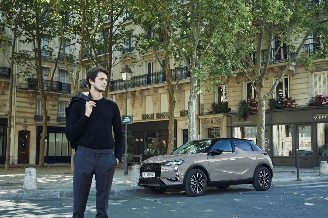 Nouvelle DS 3 grise garée dans une rue parisienne bordée d'arbres et d'immeubles haussmanniens, un homme en vêtements noirs à proximité