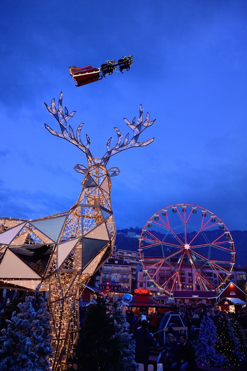 Traîneau du Père Noël survolant une grande structure de renne illuminée dans le marché de Montreux Noël, avec roue et chalets en arrière-plan