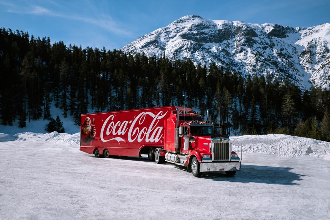 Le camion de Noël Coca-Cola rouge stationné dans un paysage alpin enneigé avec montagne et forêt en arrière-plan