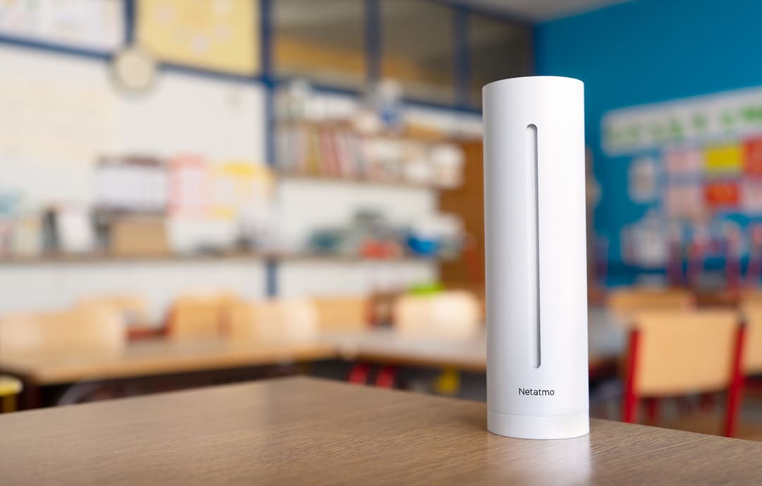 Capteur de Qualité de l'Air Intelligent Netatmo blanc posé sur un bureau en classe, arrière-plan flou avec mur turquoise et mobilier scolaire