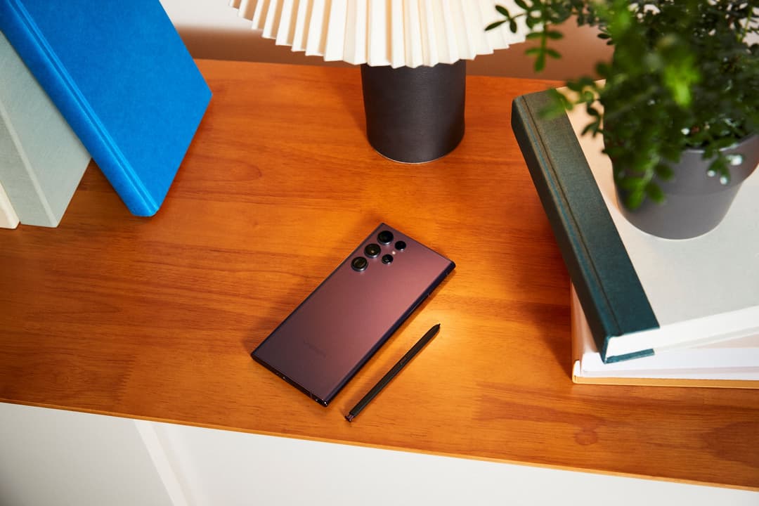 Smartphone Samsung bordeaux avec stylet posé sur un bureau en bois, entouré de livres et d'objets de décoration