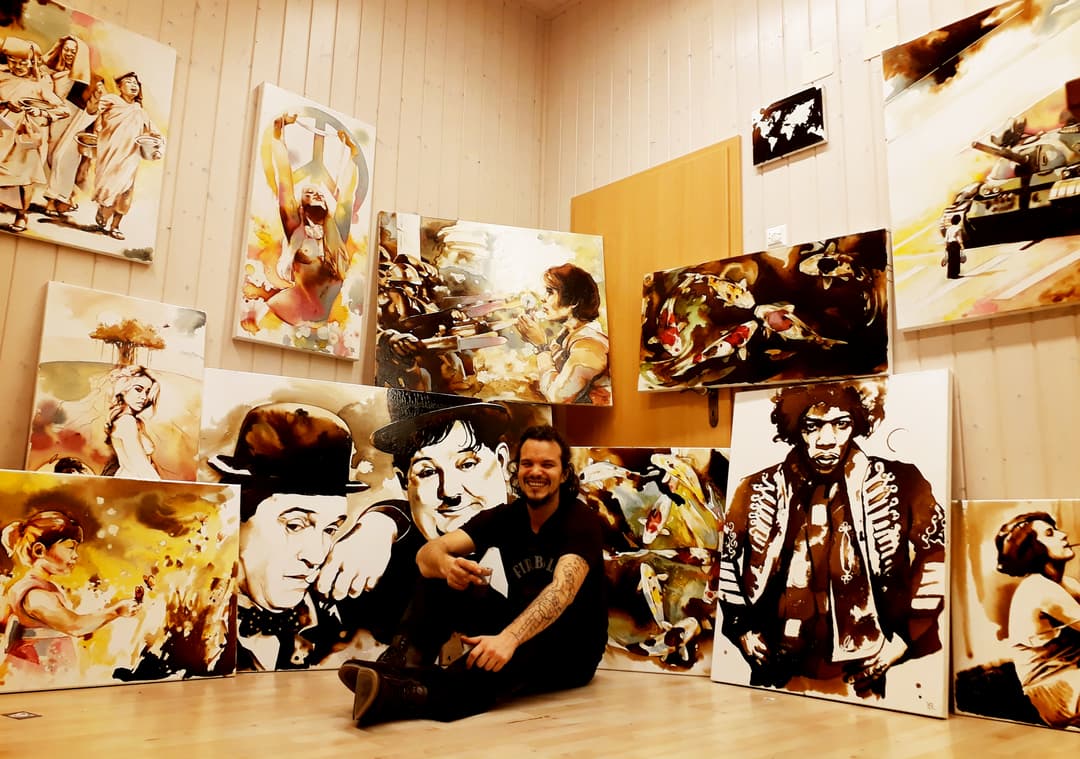 Artiste assis devant ses peintures murales aux tons sépia et or, représentant des personnages et visages stylisés