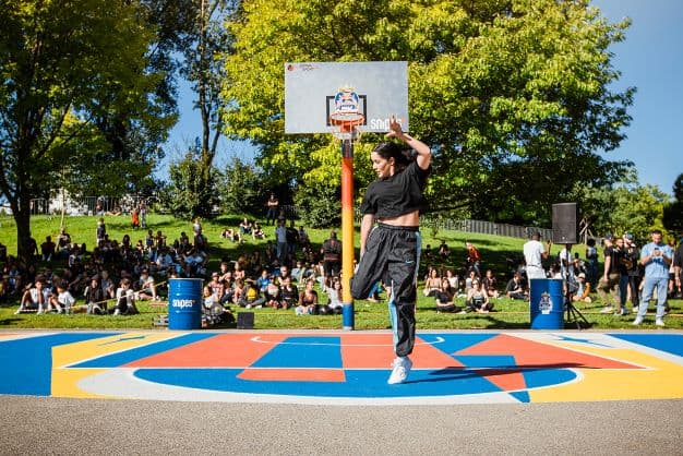 Danseur en noir exécutant un mouvement acrobatique sur un terrain de basket coloré devant une foule en plein air
