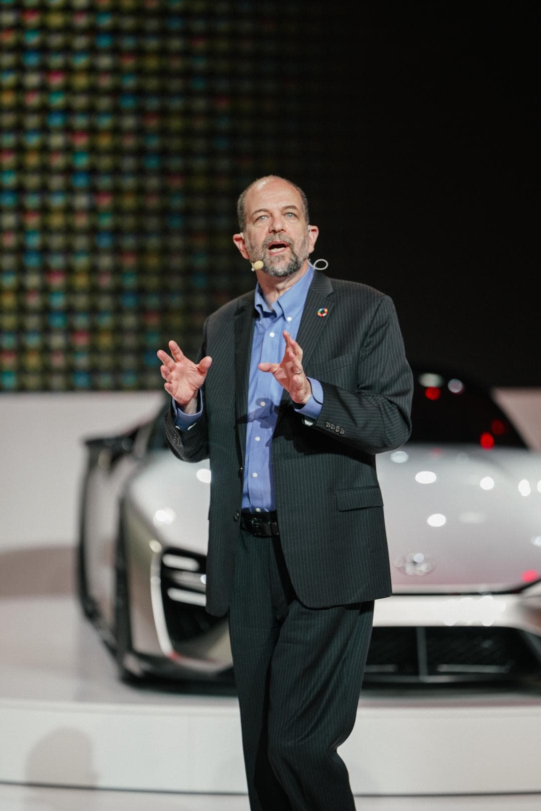 Dr Gill Pratt en costume gris clair s'exprimant au microphone lors d'une présentation, avec un véhicule Toyota en arrière-plan