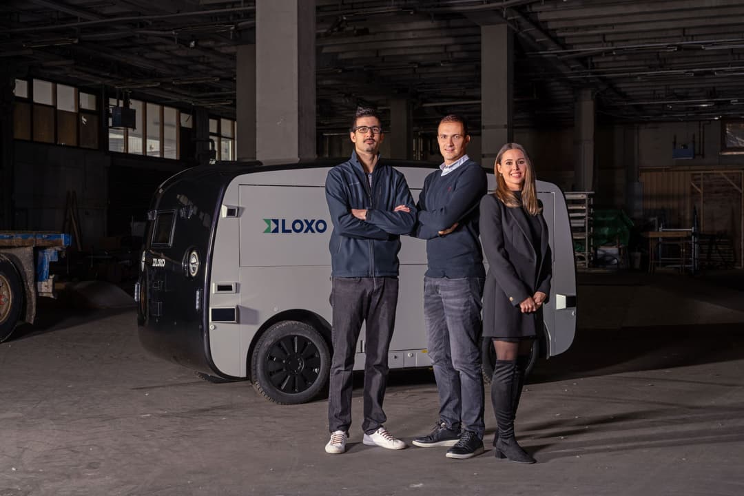 Trois personnes posent devant un véhicule autonome blanc LOXO dans un entrepôt industriel