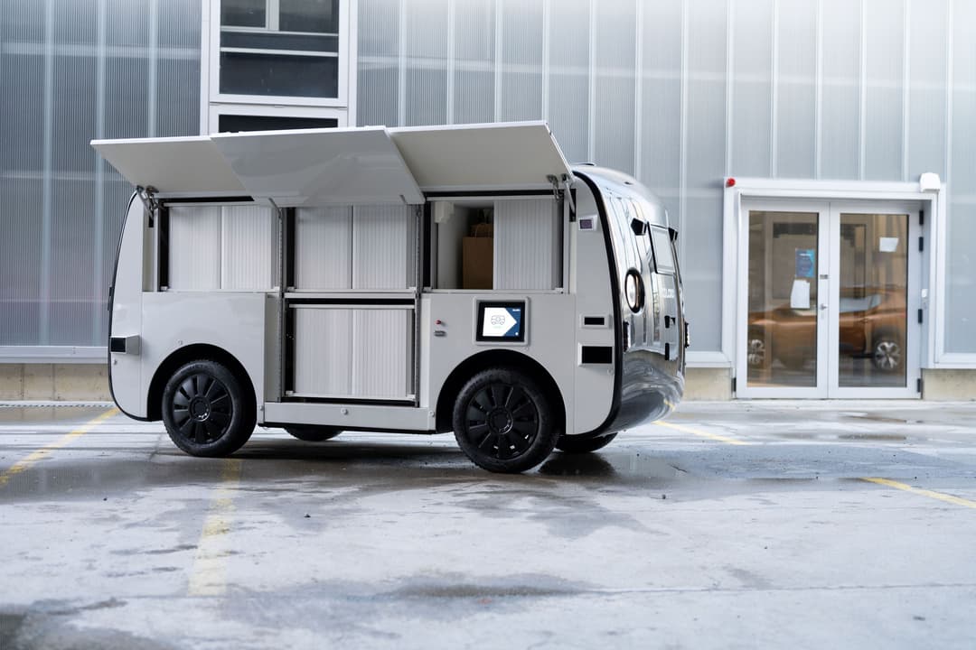 LOXO, petit véhicule de livraison autonome blanc et noir avec portes ouvertes, devant un bâtiment moderne