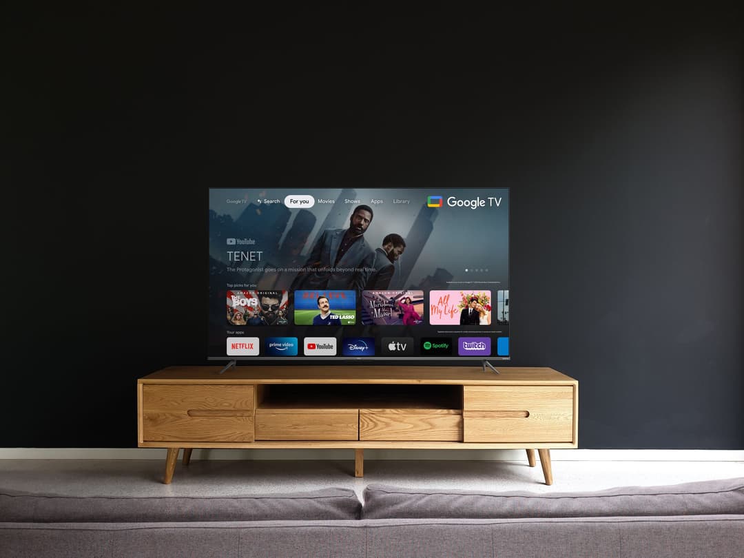 Téléviseur TCL C63 Series affichant l'interface Google TV, posé sur un meuble en bois clair dans un salon