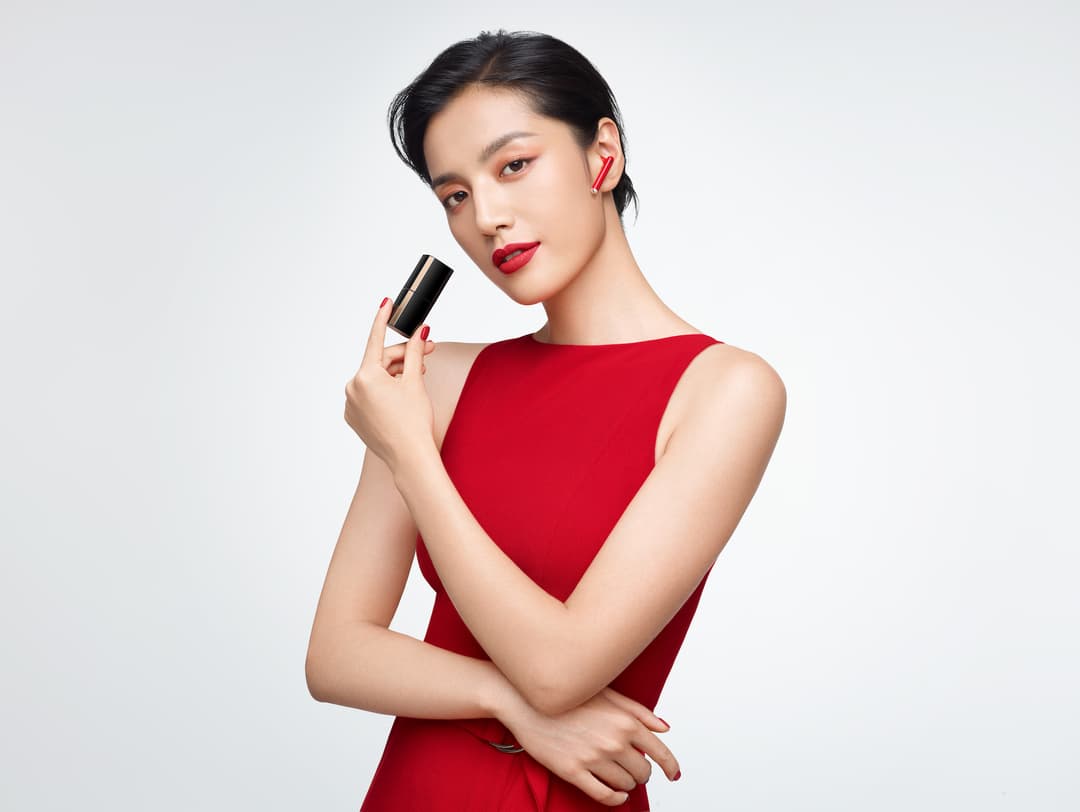 Femme en robe rouge tenant un étui noir et or en forme de rouge à lèvres, avec un écouteur Huawei FreeBuds Lipstick rouge à l'oreille