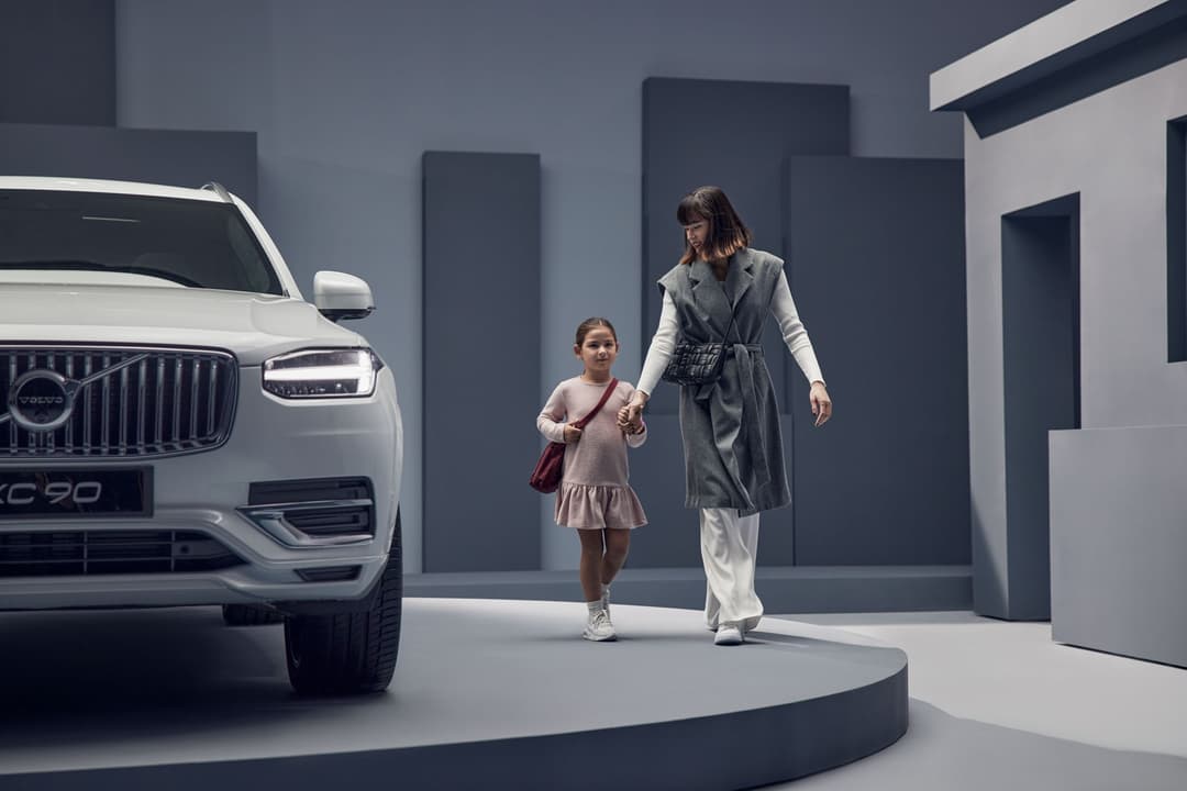 Volvo XC90 blanc avec une femme et une enfant qui marchent dans un garage moderne aux tons gris et blanc