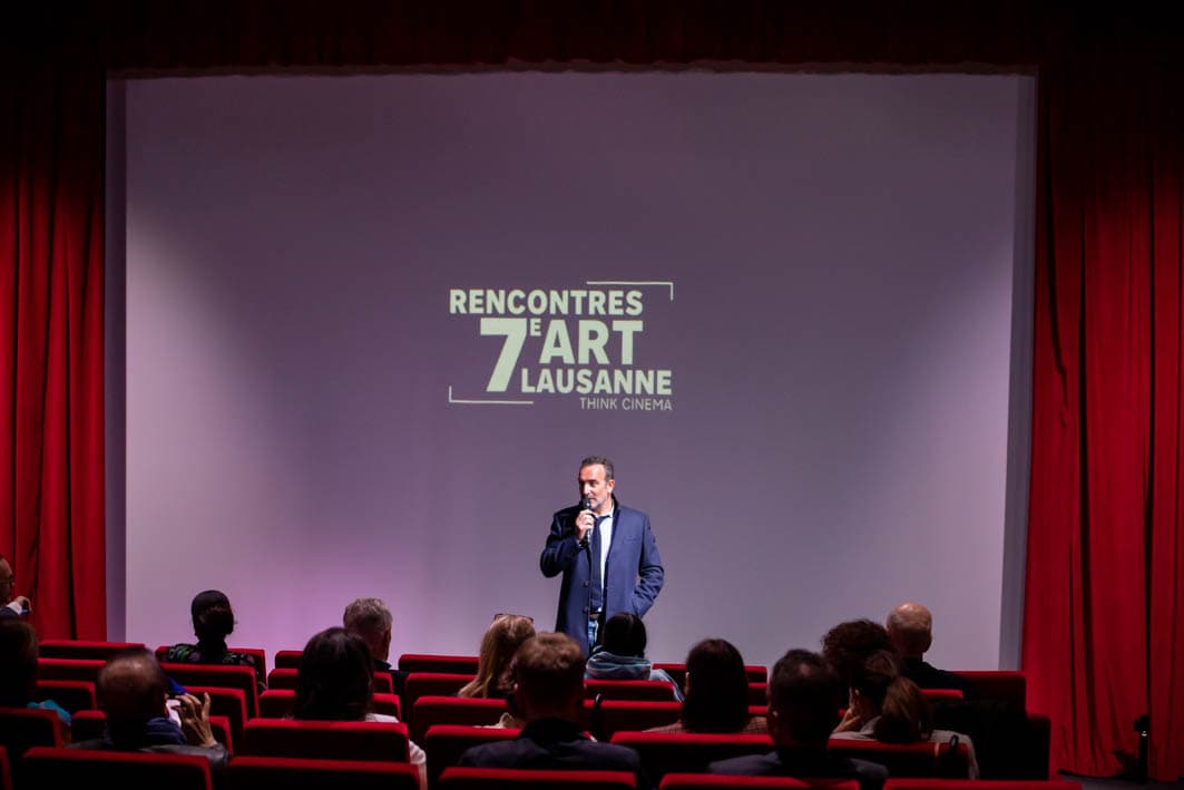 Un homme au micro sur scène dans une salle de cinéma, devant le logo des Rencontres du 7e Art Lausanne, face à un public assis dans des fauteuils rouges