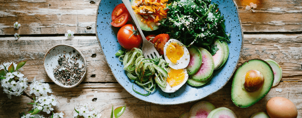 Assiette bleue remplie de légumes frais, œufs et herbes, entourée d'avocats et de fleurs blanches sur table en bois