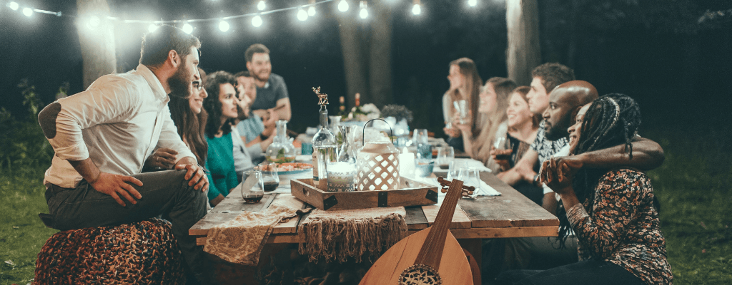 Groupe d'amis réunis autour d'une table en bois le soir, partageant un repas à la lumière des guirlandes suspendues