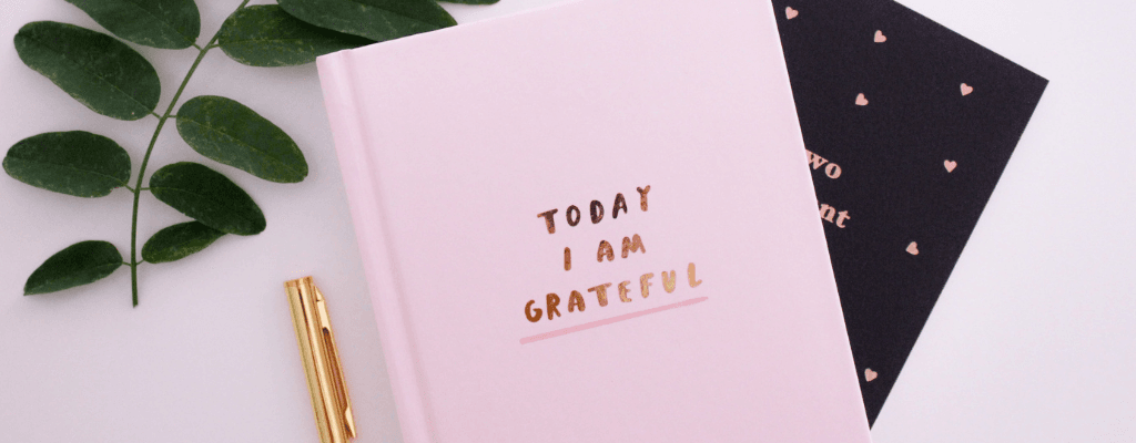 Carnet rose avec le texte « Today I am grateful » entouré d'une plante verte et d'un stylo doré