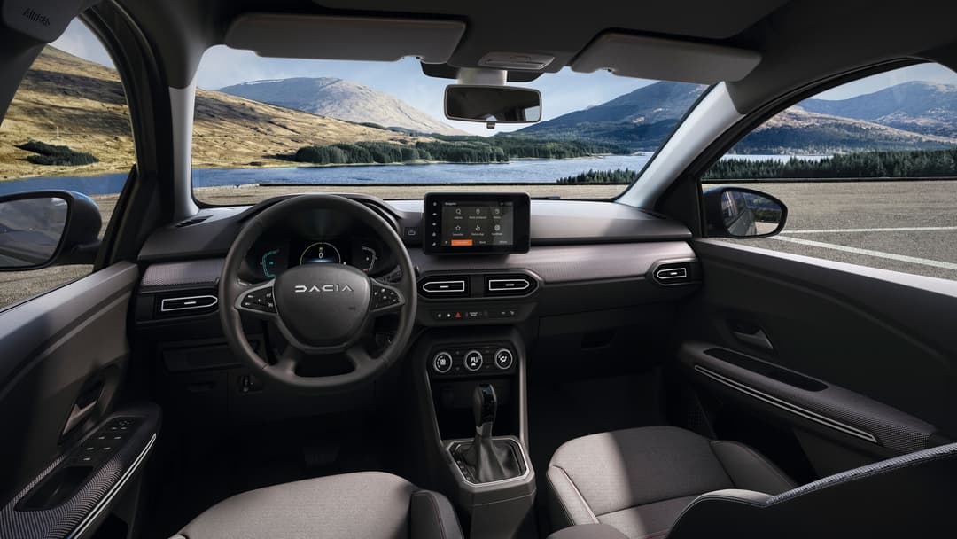 Intérieur du Dacia Jogger Hybrid 140 avec volant, écran tactile et console, vue sur montagne et lac à travers le pare-brise
