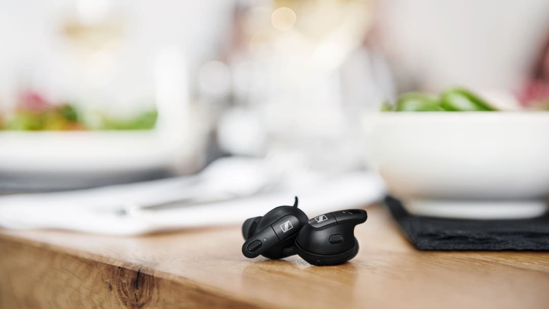 Écouteurs Sennheiser noirs posés sur un plan de travail en bois, avec des assiettes et couverts flous en arrière-plan