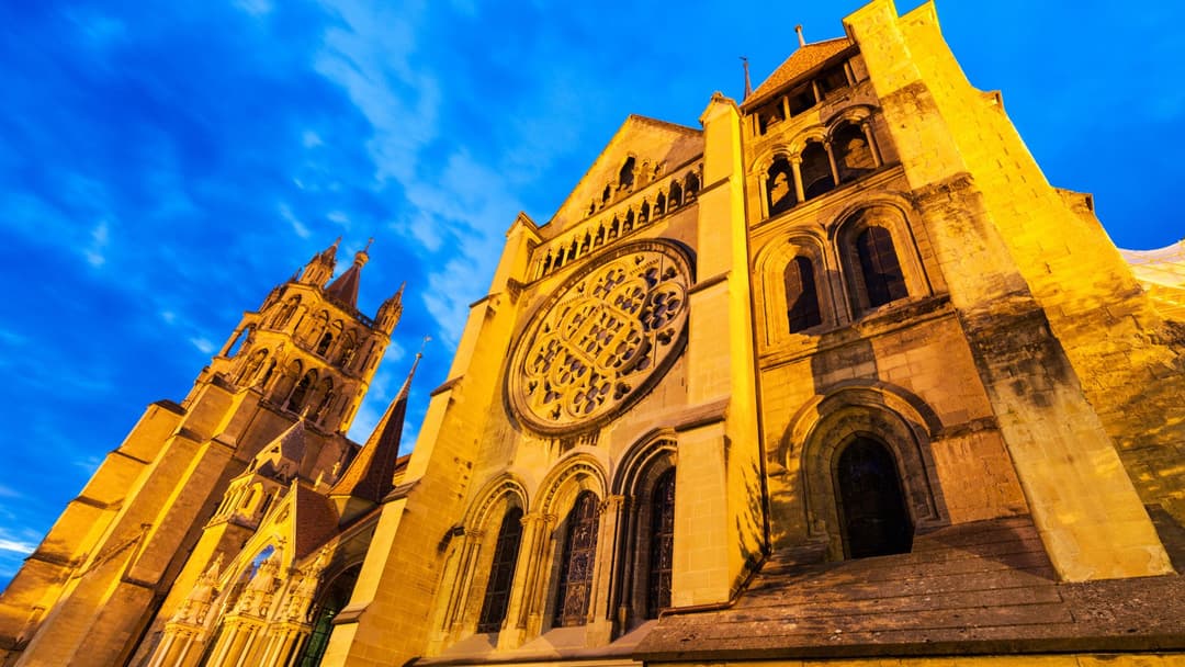 La cathédrale de Lausanne illuminée par le soleil couchant, ses façades ocre et ses tours pointues se détachant sur un ciel bleu clair