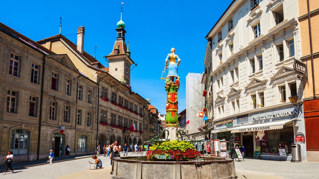 Place de la Palud à Lausanne avec sa fontaine centrale ornée d'une statue dorée, entourée de bâtiments historiques et d'une tour de l'horloge sous un ciel bleu