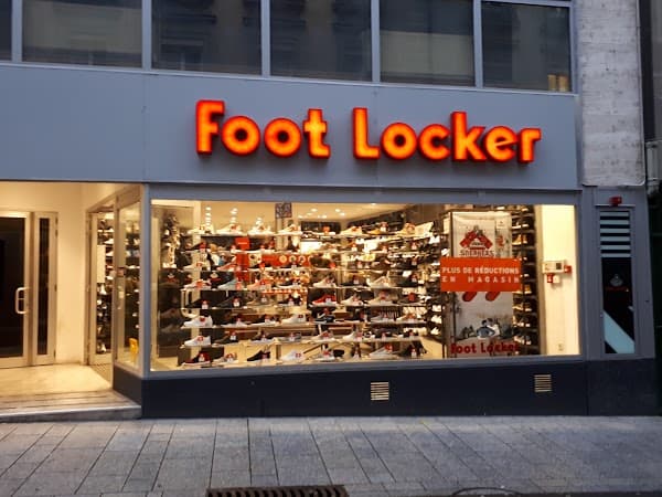 Devanture du magasin Foot Locker à Lausanne avec enseigne rouge et orange, vitrine éclairée présentant chaussures de sport et vêtements de marques Nike, Adidas, Puma et Converse