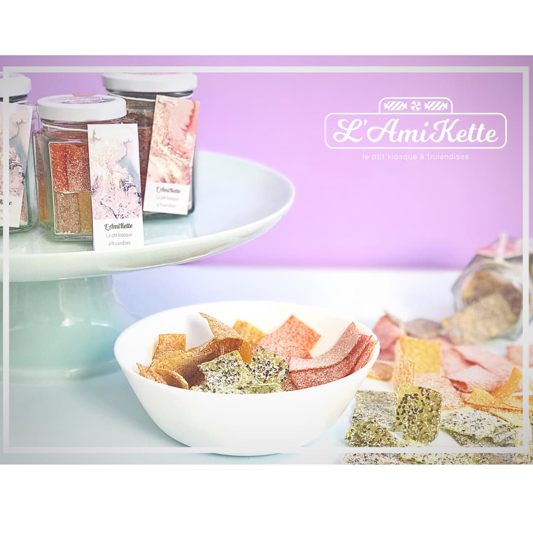 L'AmiKette, petit kiosque à friandises, présente ses bonbons artisanaux colorés dans des bocaux et une assiette blanche, sur fond rose pastel