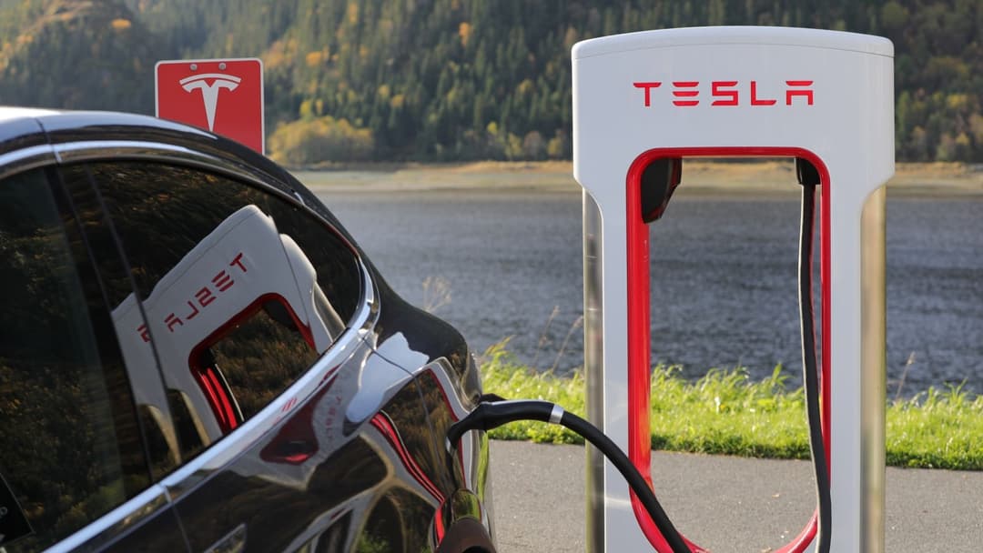 Véhicule électrique Tesla en charge sur une borne Supercharger blanche et rouge, au bord d'un lac avec forêt en arrière-plan