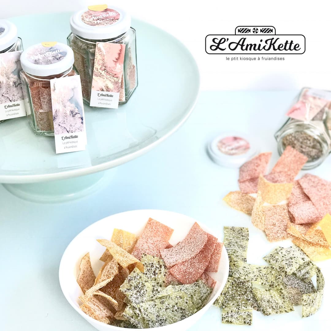Bonbons L'AmiKette en verre avec étiquettes marbre rose et blanc, présentés dans une assiette bleue pâle avec des bonbons colorés jaunes, roses et verts