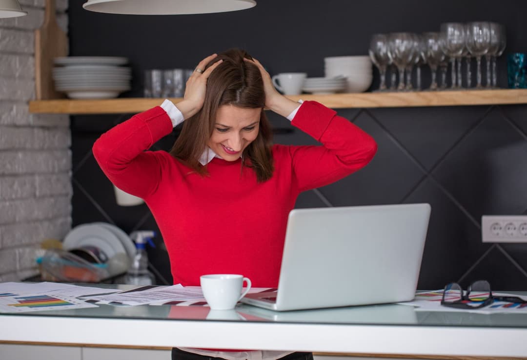 Femme en pull rouge souriant devant son ordinateur portable, les mains sur la tête, dans un espace de travail moderne