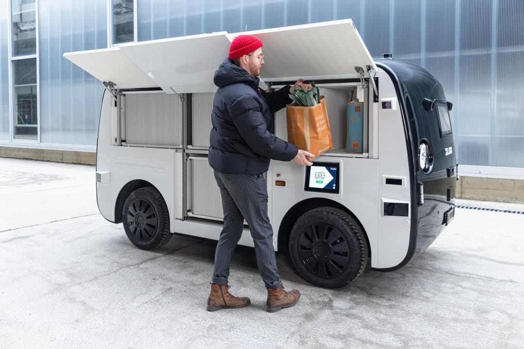 Migronomous, petit véhicule électrique blanc sans chauffeur, avec un homme prélevant un sac de courses Migros dans son compartiment ouvert