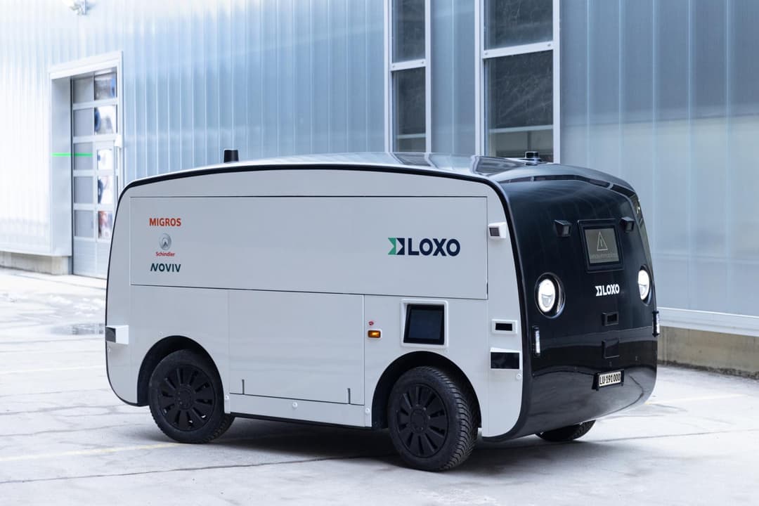 Véhicule de livraison autonome Loxo blanc et noir aux logos Migros, Schindler et Noviv, stationné devant un bâtiment moderne
