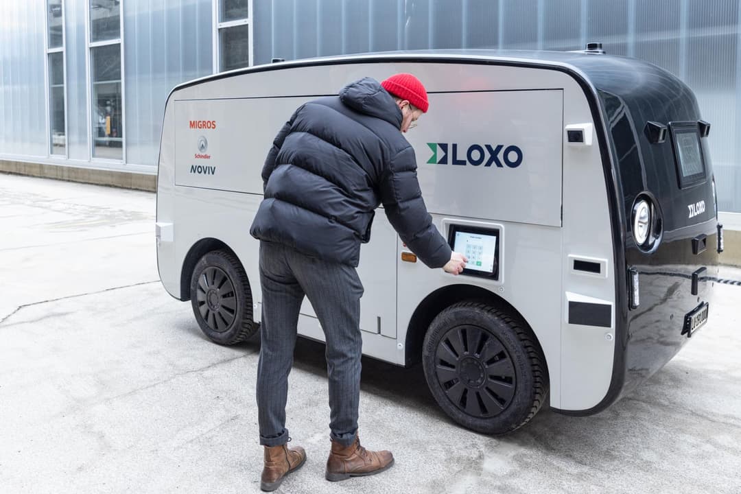 Un homme avec un bonnet rouge interagit avec un petit véhicule autonome blanc Loxo aux couleurs de Migros, équipé d'un écran tactile de livraison