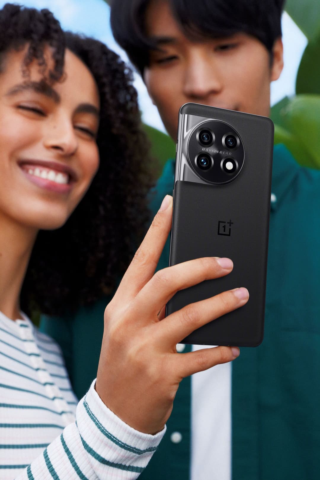 OnePlus 11 noir tenu en main avec deux personnes en arrière-plan souriantes, mettant en avant le module photo circulaire du smartphone