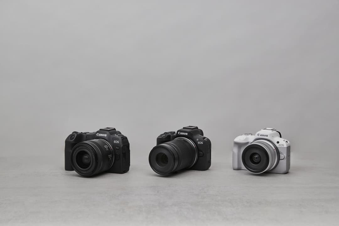 Trois boîtiers Canon EOS R50 alignés : deux noirs et un blanc, équipés d'objectifs, sur fond gris