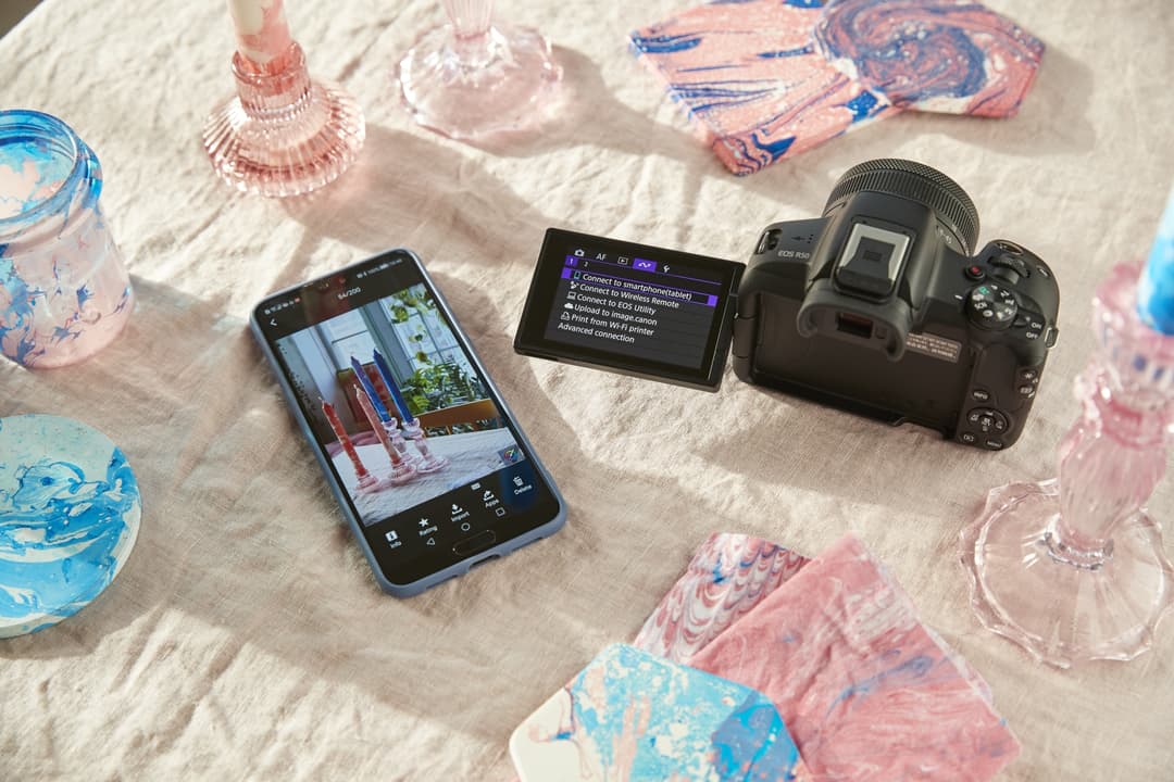 Canon EOS R50 noir avec écran tactile affichant les paramètres, à côté d'un smartphone, sur un tissu blanc avec accessoires colorés