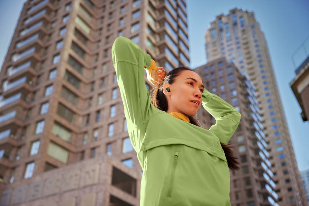 Femme portant un casque audio Philips GO Sport noir en extérieur, vêtue d'une veste vert fluo et d'un foulard orange, devant des immeubles urbains