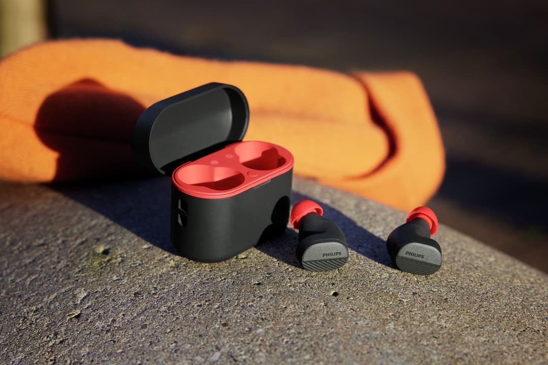 Écouteurs sans fil Philips GO Sport TAA5508 noir et rouge avec boîtier de charge ouvert, posés sur du sable avec un sac orange en arrière-plan