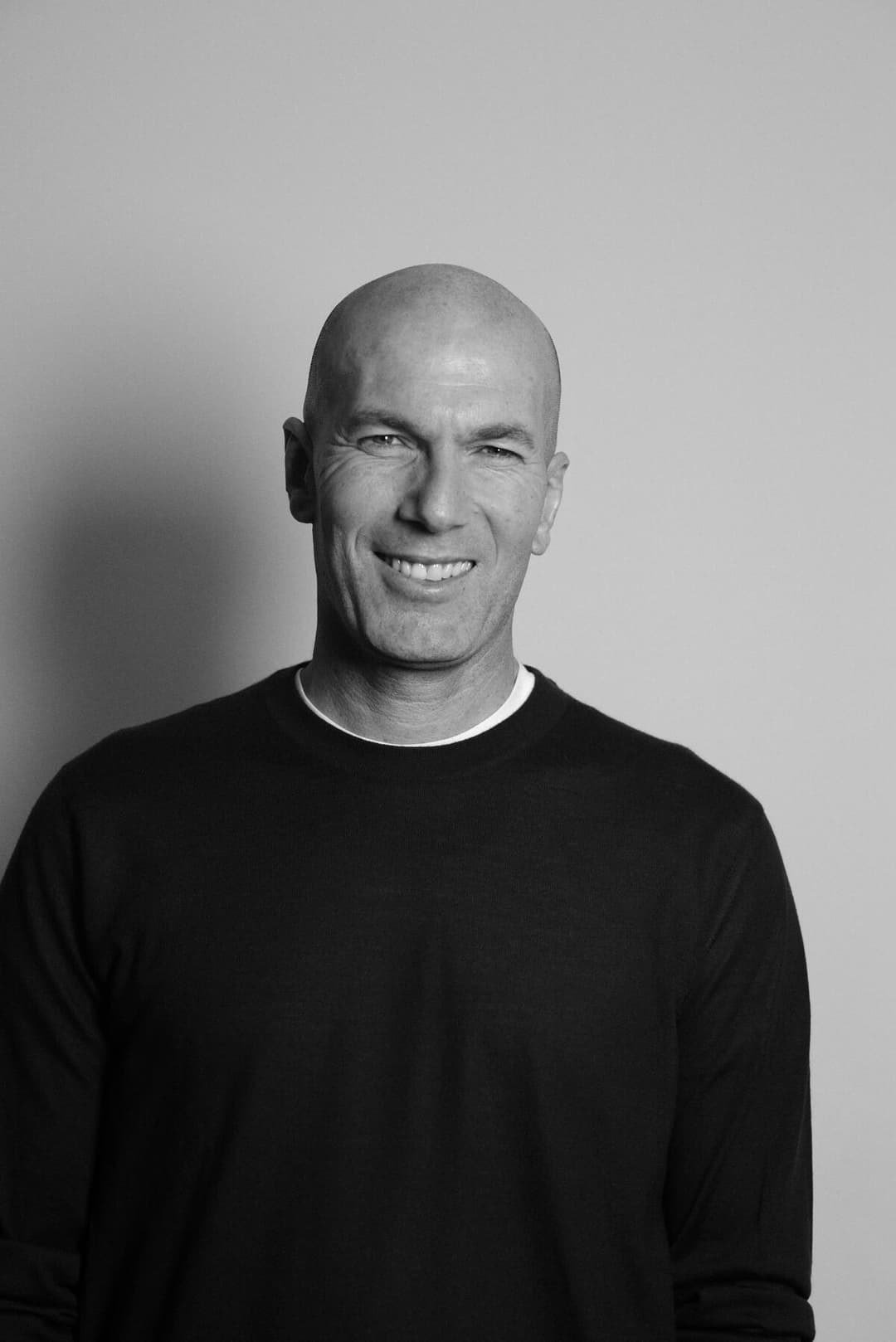 Zinedine Zidane en portrait noir et blanc, souriant, vêtu d'un pull noir col rond