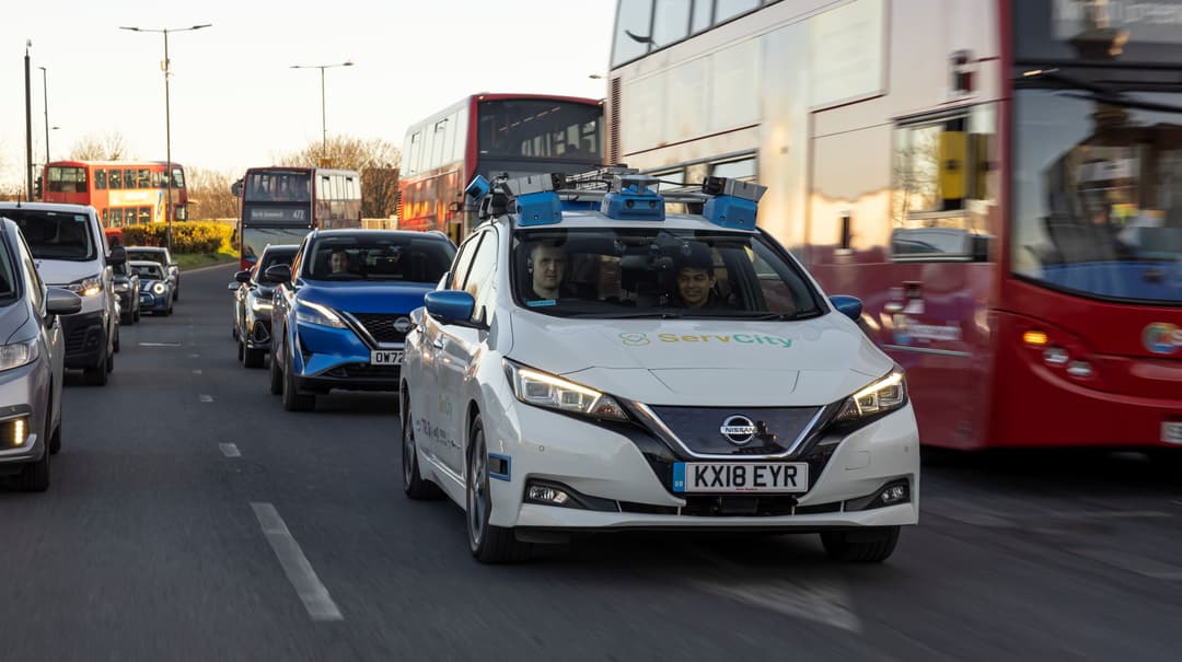 Nissan LEAF blanche équipée de capteurs autonomes en circulation urbaine dense, entourée de bus et d'autres véhicules